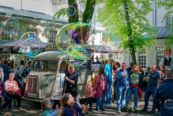 Seifenblasenwalkact mit Riesenseifenblasen Menschen feiern auf einer Straße, umgeben von Luftballons und einem alten Lastwagen.