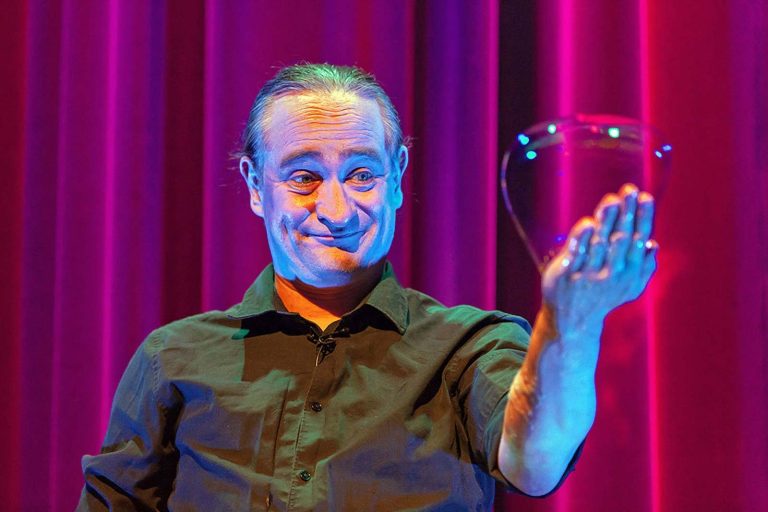 Seifenblasenkünstler Stephan Masur Handseifenblase Lächelnder Mann in schwarzem Hemd hält eine klare Glasvase vor einem rosanen Hintergrund.