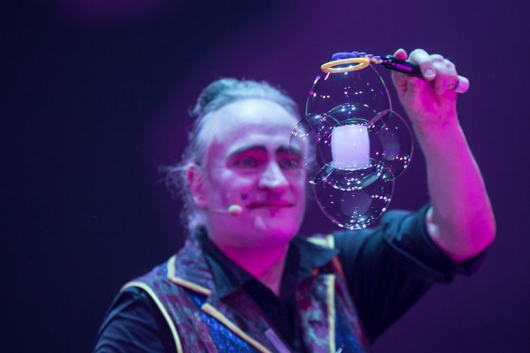 Seifenblasenshow von Stephan Masur Ein Zirkuskünstler hält eine große Seifenblase vor einem bunten Hintergrund.