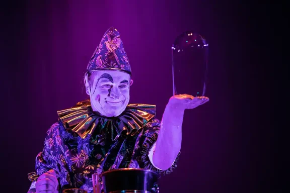 Seifenblasenclown Stephan Masur Clown in bunting costume hält ein Glasgefäß, mit lila Hintergrundbeleuchtung.