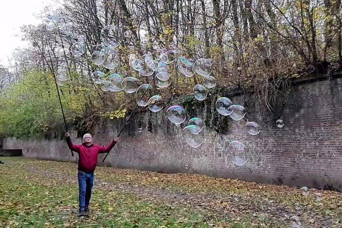 Mann in rotem Sweatshirt steht im Park und bläst große Seifenblasen.